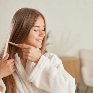young woman smiles and enjoys her beauty, combes long straight hair after applying mask, stands