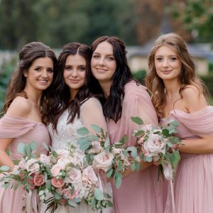 Beautiful bride with bridesmaids dressed in pink dresses are holding pale pink bouquets made of roses outdoors