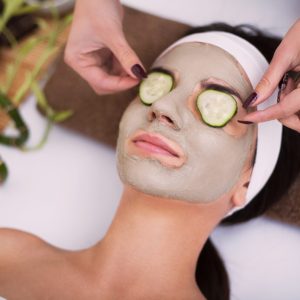 Spa. Beautiful young woman is getting facial clay mask at spa, lying with cucumbers on eyes